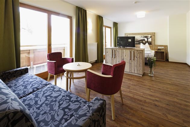 Ein gemütliches Zimmer mit einem Wohnzimmerbereich, einem Tisch und zwei Stühlen. Große Fenster lassen viel Licht herein und bieten einen Blick auf den Balkon.