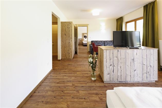 Ein modernes Zimmer mit Holzfußboden und einer gemütlichen Einrichtung. Der Raum verfügt über einen Fernseher und große Fenster mit grünen Vorhängen.