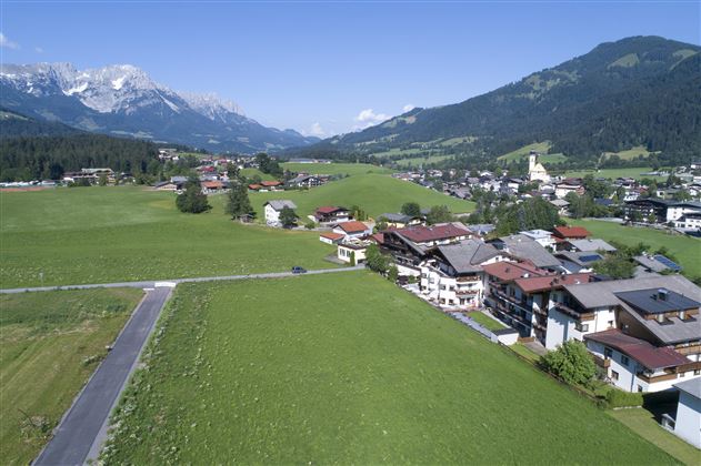 Eine malerische Landschaft mit grünen Wiesen und kleinen Häusern. Im Hintergrund sind Berge und ein klarer Himmel zu sehen.