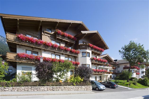 Ein charmantes Gebäude im alpinen Stil mit bunten Blumenbalkonen. Die Umgebung ist grün und einladend, ideal für einen entspannten Aufenthalt.