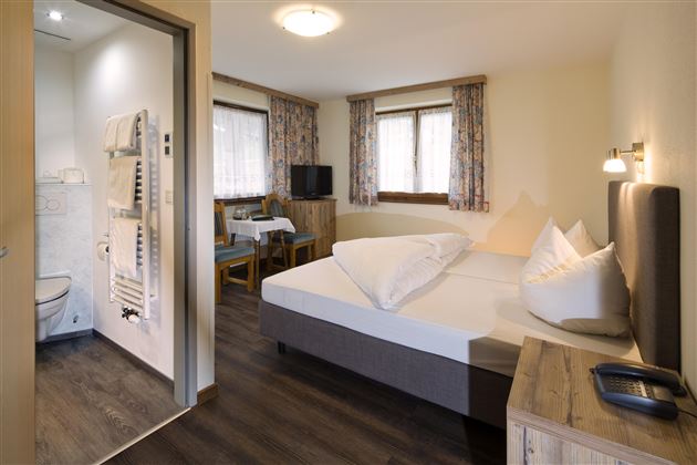 Ein gemütliches Schlafzimmer mit einem Doppelbett und zwei Kissen. Im Hintergrund sind ein Tisch, Stühle und ein Badezimmer sichtbar.