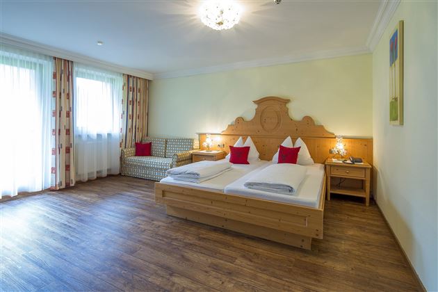 Ein gemütliches Hotelzimmer mit einem Doppelbett und zwei roten Kissen. Große Fenster sorgen für viel Tageslicht und eine angenehme Atmosphäre.