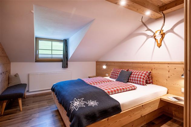 Een gezellige slaapkamer met houten panelen en een groot bed. Aan de wand hangt een geweer, en het raam laat veel daglicht binnen.