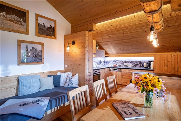 Een gezellige woonkeuken met houten wandbekleding en lichte muren. Op de tafel staat een vaas met zonnebloemen en er is een uitzicht op de bergen door het raam.