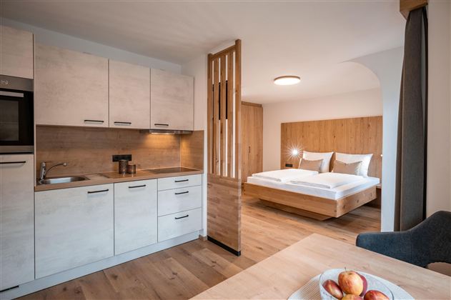 Světlý pokoj s otevřenou kuchyní a pohodlnou postelí. Moderní design a dřevěné akcenty vytvářejí útulnou atmosféru.