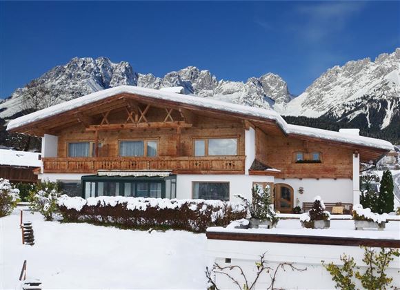 A beautiful chalet in the snow, surrounded by majestic mountains. The clear blue sky gives the winter landscape a serene atmosphere.