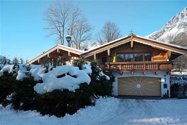 Ein schönes Holzhaus im Schnee mit einem großen Balkon. Umgeben von verschneiten Bäumen und einem klaren blauen Himmel.