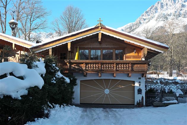 Ein charmantes Holzhaus mit einem Balkon, umgeben von schneebedeckten Bäumen. Im Hintergrund sind Berge unter einem klaren blauen Himmel sichtbar.