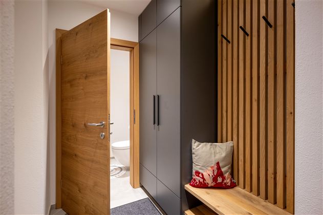 Ein moderner Eingangsbereich mit einer Holz- und grauen Schrankkombination. Im Hintergrund ist eine geöffnete Tür zu einer Toilette sichtbar.