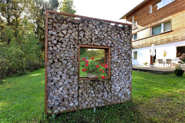 A creatively designed woodpile with a window through which a flower is visible. In the background, there is a modern house and a green meadow.