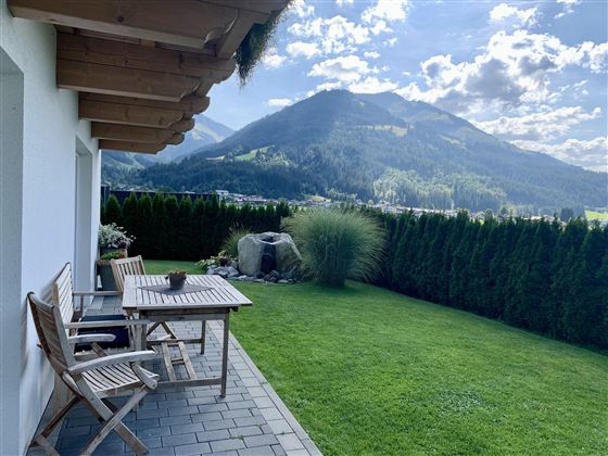 Ein schöner Garten mit Rasen und einer Terrasse. Im Hintergrund sind majestätische Berge und ein blauer Himmel zu sehen.