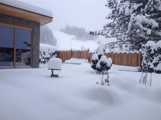 A snow-covered landscape with a wooden cabin and snow-covered trees. The snow lies thick on the ground and covers the garden furniture.