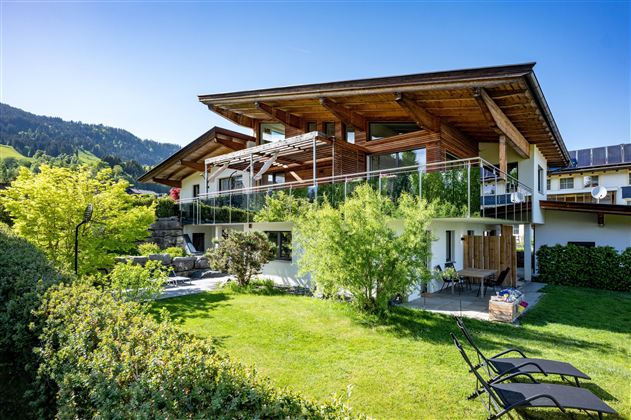 A modern house with a spacious garden and terrace. The surroundings are green and surrounded by mountains.