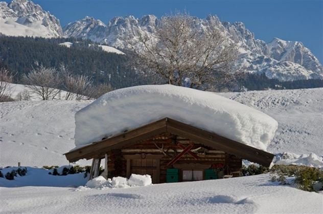 Eine winterliche Landschaft mit einem kleinen Holzhaus, das unter einer dicken Schneeschicht verborgen ist. Im Hintergrund sind hohe Berge und ein klarer blauer Himmel sichtbar.
