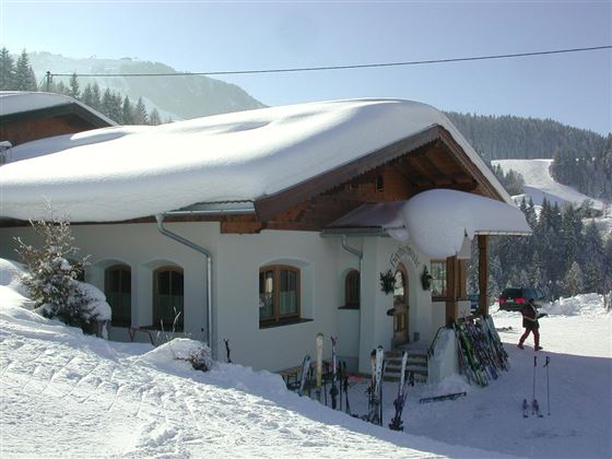 Ein charmantes Haus im Schnee mit einem schneebedeckten Dach. Im Vordergrund stehen Skier, und die Umgebung ist winterlich und idyllisch.