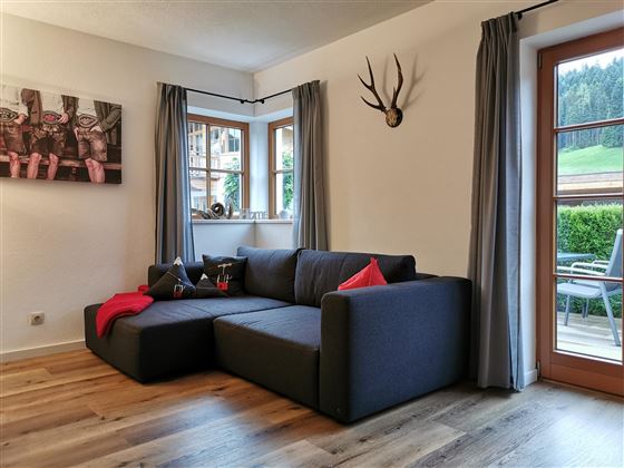Ein moderner Wohnraum mit einem bequemen, grauen Sofa und einem roten Kissen. Große Fenster bieten einen Blick ins Freie und die Wände sind dezent dekoriert.