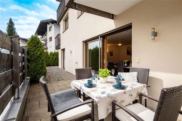 Ein gemütliche Sonnenterrasse mit Tisch und Stühlen, gedeckt für Kaffee und Kuchen. Im Hintergrund ist eine grüne Hecke und ein modernes Gebäude zu sehen.