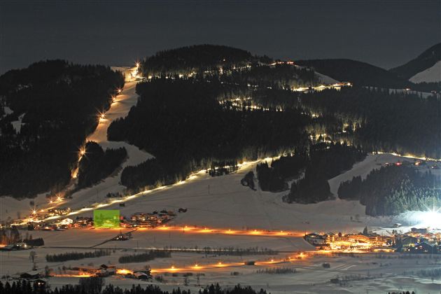 Een besneeuwd landschap bij nacht met verlichte skipistes en kleine huisjes. De lucht is donker en de lichten van het dorp verlichten de scène.