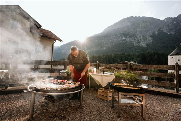 Ein Grillmeister bereitet verschiedene Speisen im Freien zu, umgeben von einer malerischen Berglandschaft. Der Rauch steigt in die Luft, während die Sonne im Hintergrund scheint.