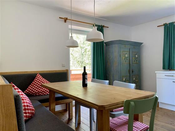 A cozy dining area with a wooden table and colorful chairs. In front of the window are green curtains and a bottle of wine on the table.