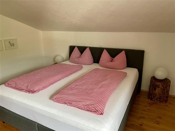 A cozy bedroom with a double bed and plaid bedspreads. The walls are bright, and a wooden chair and a lamp complement the decor.