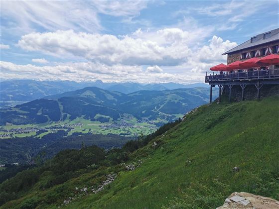 Eine Berglandschaft mit einem Restaurant, das über eine Terrasse mit roten Schirmen verfügt. Die grünen Wiesen und das weitläufige Tal bieten einen schönen Blick auf die umliegenden Berge.