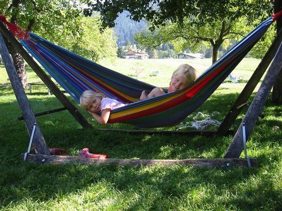 Twee kinderen liggen in een kleurrijke hangmat onder een boom. De omgeving is groen en zonnig, met uitzicht op een weide op de achtergrond.