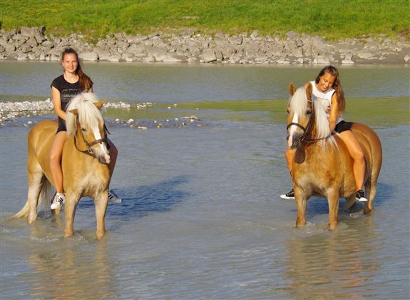 Two girls are riding horses in the shallow water. In the background, a green landscape can be seen.