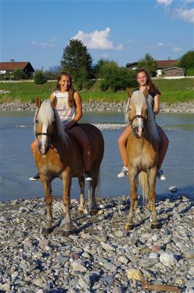 Two girls are riding horses by the riverbank. In the background, green trees and houses are visible.