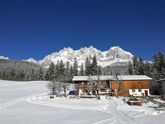 Eine malerische Winterlandschaft mit einem Holzhaus und schneebedeckten Bergen im Hintergrund. Der strahlend blaue Himmel sorgt für eine friedliche Atmosphäre.