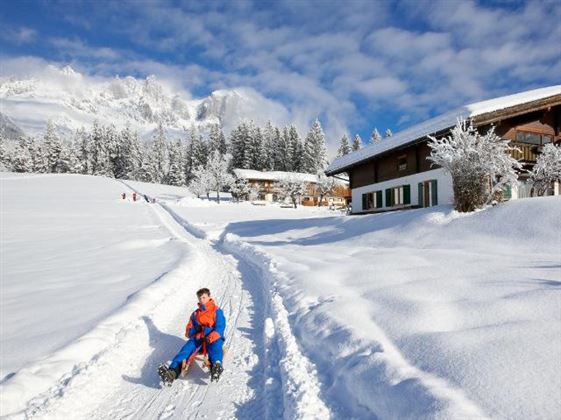 Ein Junge sitzt auf einem Schlitten in einer schneebedeckten Landschaft. Im Hintergrund sind schneebedeckte Bäume und gemütliche Häuser zu sehen.