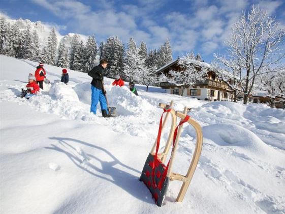 Eine verschneite Winterlandschaft mit mehreren Menschen, die Schnee schaufeln. Im Vordergrund steht ein Schlitten, während im Hintergrund ein Haus und schneebedeckte Bäume sichtbar sind.