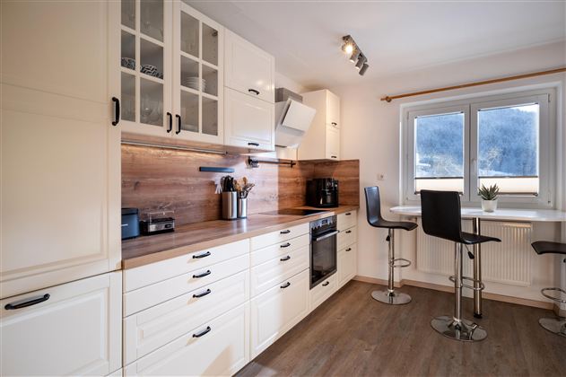 Een moderne keuken met witte kasten en houten oppervlakken. Er is een kleine eethoek met hoge stoelen en veel daglicht.