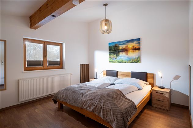 Een gezellige slaapkamer met een groot bed en een houten standaard. Aan de muur hangt een afbeelding van een landschap, en het licht valt door de ramen.