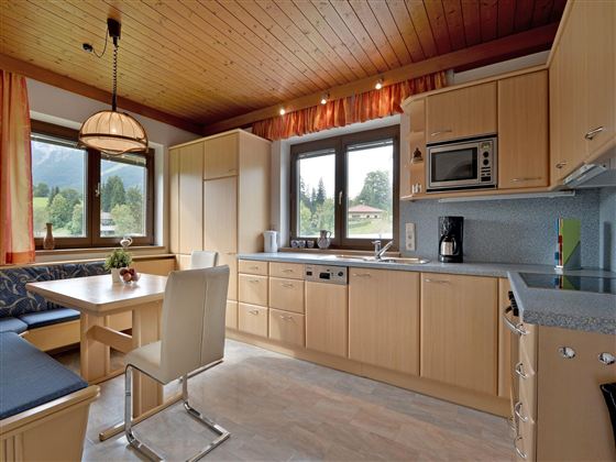Eine moderne Küche mit Holzdecke und großen Fenstern. Der Essbereich hat einen Tisch und Stühle, sowie eine Küchenzeile mit verschiedenen Geräten.