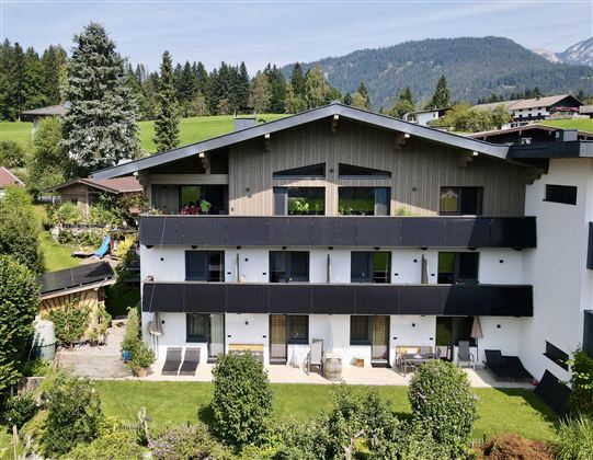 A modern building surrounded by green meadows and trees. In the background, mountains and a blue sky can be seen.