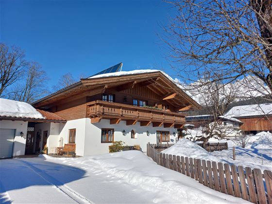 A traditional chalet in winter with a snow-covered plot. The blue sky and the surrounding trees give the scene a serene atmosphere.