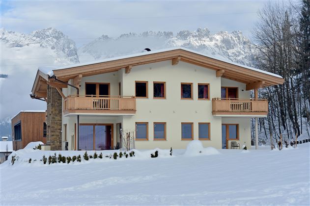 Ein modernes Haus im Schnee mit Balkon und großen Fenstern. Im Hintergrund sind schneebedeckte Berge zu sehen.