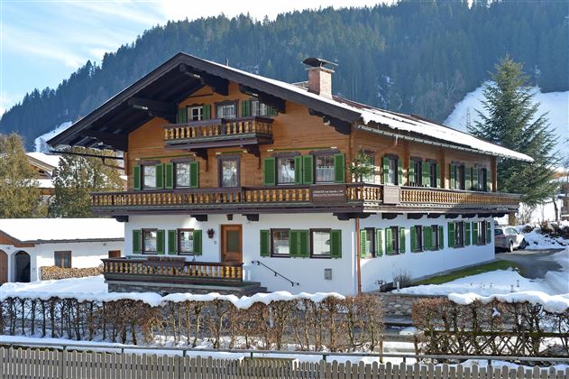 A traditional alpine house with a wooden balcony and green shutters. The garden is surrounded by snow-covered areas and wooded hills can be seen in the background.