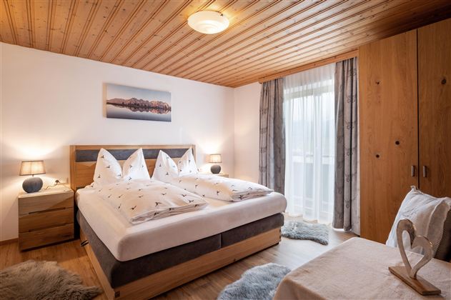 A cozy bedroom with a large bed and decorative pillows. The wooden details and the window create a warm atmosphere.
