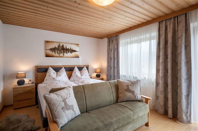 A cozy bedroom with a double bed and two nightstands. Large windows and a sofa create an inviting atmosphere.