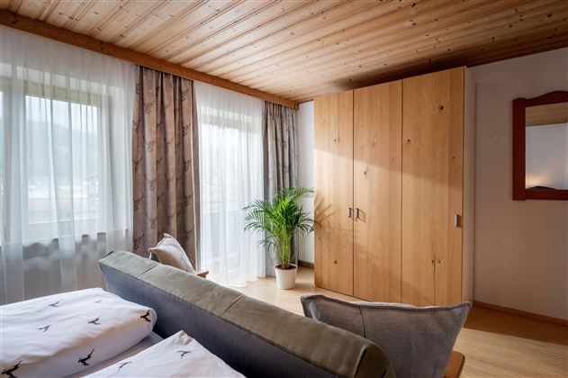 A cozy bedroom with a wooden ceiling and bright curtains. The room is decorated with a large wardrobe and a plant.