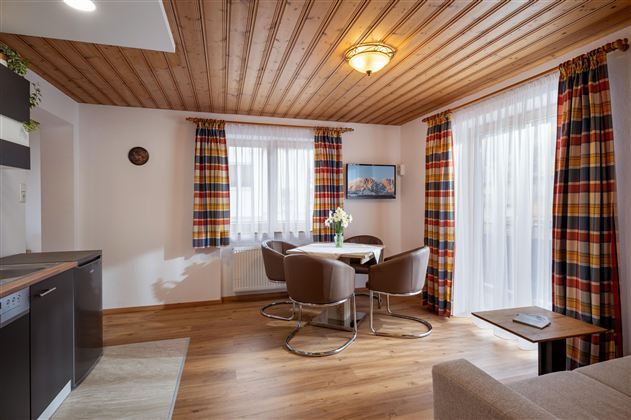 A cozy living space with a wooden ceiling, large window front, and colorful curtains. A dining table with modern chairs and a small kitchen corner are also visible.
