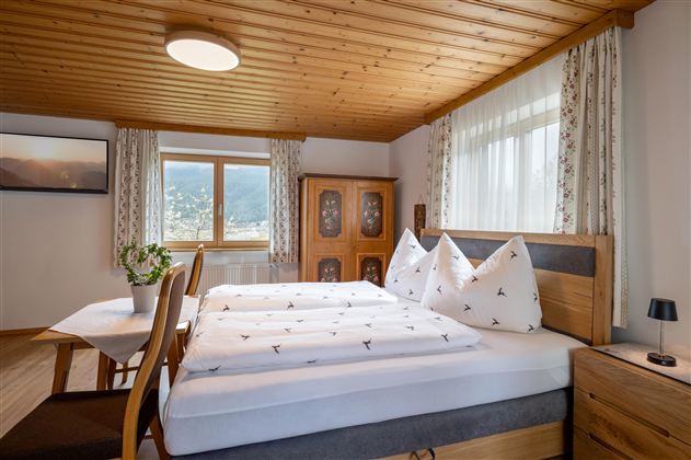 A cozy bedroom with a large bed and wooden details. Large windows provide plenty of daylight and a pleasant atmosphere.