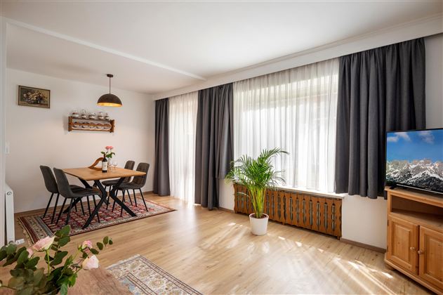 A bright living room with a dining table, chairs, and a large window front. Plants and a television give the room a cozy atmosphere.