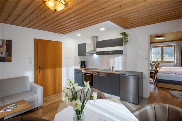 A modern kitchen with a dining area and a cozy living space. The bright room has windows overlooking the outside and a wooden ceiling.