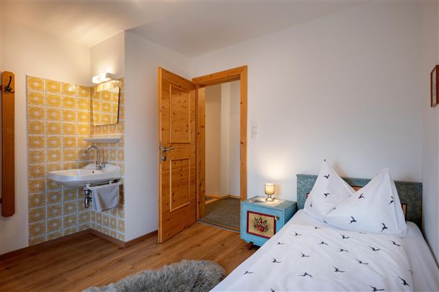 A cozy bedroom with a single bed and hardwood floor. The walls are painted light, and there is a small sink as well as a door to another room.