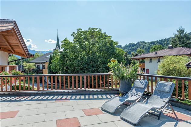 A terrace with two loungers and a view of a green landscape. In the background, houses and trees can be seen.