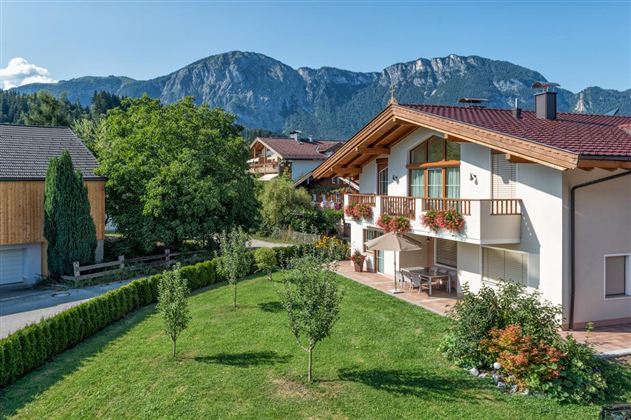A beautiful house with a balcony and flower boxes. In the background, impressive mountains and a green meadow can be seen.