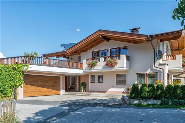 A modern house with a wooden veranda balcony and a garage. The surroundings are green with plants and a clear blue sky.